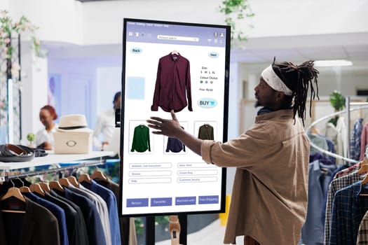 African american client selects clothes on touch screen board in clothing store, buying items from self service checkout kiosk. E commerce self ordering concept in fashion boutique.