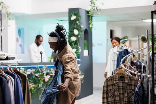 Cheerful african american man in headphones holding shirt on hanger while shopping in clothing store. Customer in good mood listening to music while choosing new outfit in fashion boutique