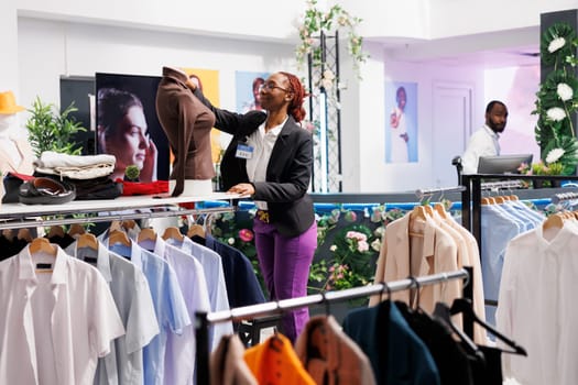 Clothing store employee putting casual blouse on mannequin to show new garment collection. Fashion boutique young african american woman assistant dressing dummy model in shop