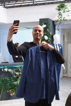 Cheerful arab man taking selfie and showcasing outfit using smartphone front camera while shopping in boutique. Blogger holding jacket and live streaming on mobile phone in clothing store