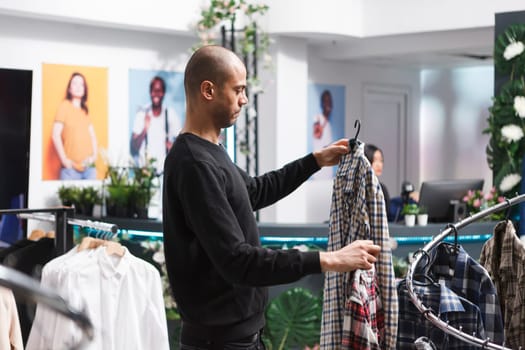 Clothing store arab customer choosing between two plaid shirts while browsing casual apparel rack. Young man standing in shopping mall boutique and examining outfit options