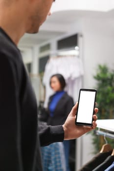 Clothing store customer holding smartphone with blank screen mockup while exploring apparel online. Boutique man hand using mobile phone with empty white display while shopping in mall