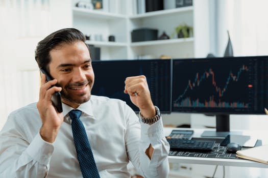 Successful in smiling trader businessman raising fist up, calling on phone against stock exchange investment analysis screen to invest high profit financial technology market at home office. Surmise.