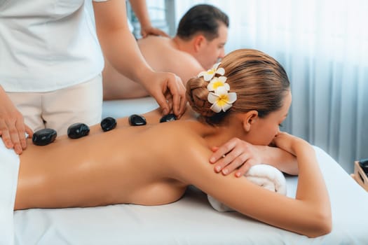 Hot stone massage at spa salon in luxury resort with day light serenity ambient, blissful couple customer enjoying spa basalt stone massage glide over body with soothing warmth. Quiescent
