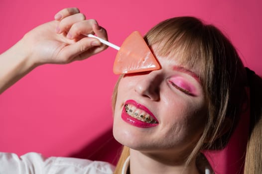 Portrait of a young woman with braces and bright makeup eating a lollipop on a pink background