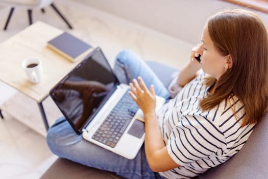 A woman multitasks by making a phone call and working on her laptop while seated on a couch with a fresh cup of coffee.