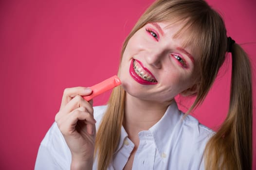 Portrait of a young woman with braces and bright makeup chewing gum on a pink background