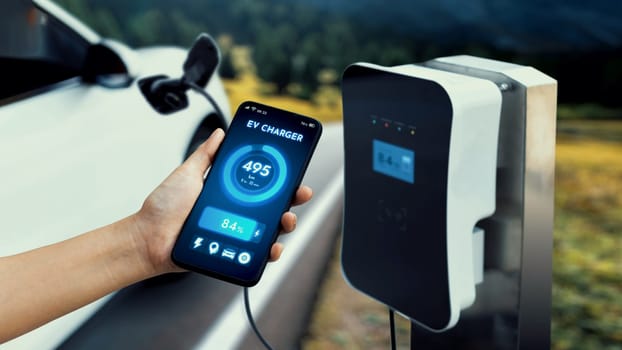 Businessman holding smartphone display battery status interface by smart EV mobile application while EV car recharging electricity from charging station with natural mountain outdoor background.Peruse