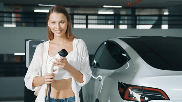 Young woman happy and smile portrait travel with EV electric car to shopp center parking lot in downtown city urban sustainability lifestyle holding EV charger plug for electric vehicle innards