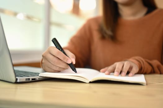 Cropped shot of young woman making important notes and using laptop on wooden desk.