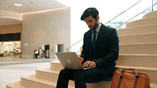 Skilled investor working or planing strategy by using laptop at stair. Professional business man wearing suit while working and typing data analysis by using laptop at modern hotel. Exultant.