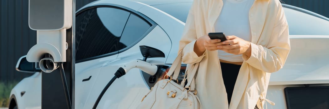 Woman holding shopping bag and use smartphone to pay for electricity to recharge EV car battery from charging station at city mall parking lot. Modern woman go shopping by eco car.Panorama Expedient