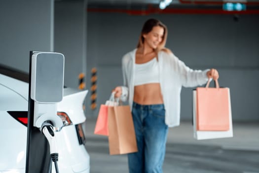 Young woman travel with EV electric car to shopping center parking lot charging in downtown city showing urban sustainability lifestyle by green clean rechargeable energy of electric vehicle innards