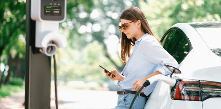 Young woman using smartphone online banking application to pay for electric car battery charging from EV charging station during vacation road trip at national park or summer forest. Panorama Exalt