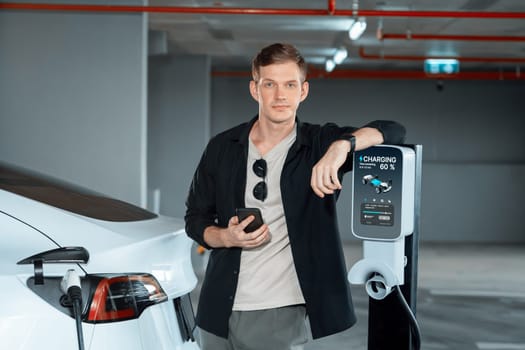 Young man travel with EV electric car to shopping center parking lot charging in downtown city showing urban sustainability lifestyle by green clean rechargeable energy of electric vehicle innards