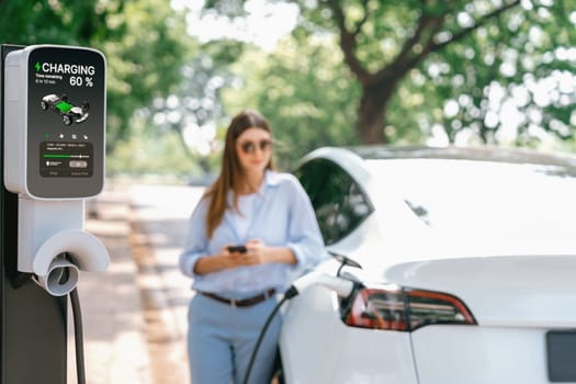 Young woman using smartphone online banking application to pay for electric car battery charging from EV charging station during vacation holiday road trip at national park or summer forest. Exalt