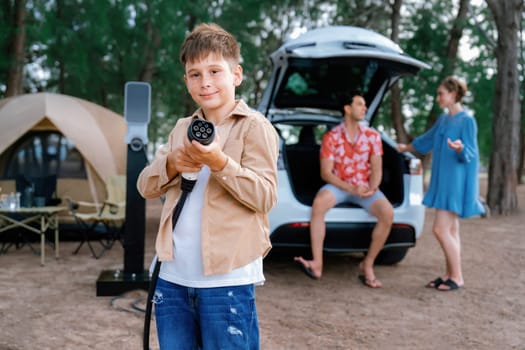 Little boy holding EV charger and point at camera with his family sitting on the trunk in background. Road trip travel with alternative energy charging station for eco-friendly car concept. Perpetual