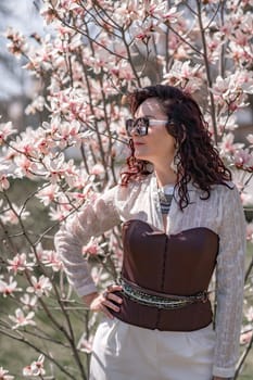 Magnolia park woman. Stylish woman in a hat stands near the magnolia bush in the park. Dressed in white corset pants and posing for the camera