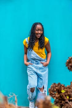 Vertical portrait of a cool young african woman leaning in a blue background wall and smiling