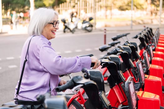 smiling senior woman taking rental bike from parking row, concept of sustainable mobility and active lifestyle in elderly people