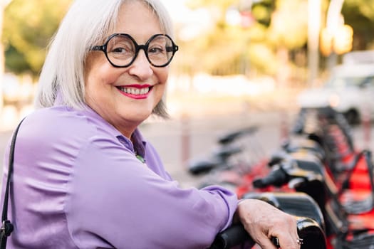 portrait of a smiling senior woman leaning on a rental bike in the parking row, concept of sustainable mobility and active lifestyle in elderly people, copy space for text