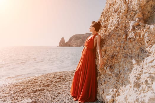 Side view a Young beautiful sensual woman in a red long dress posing on a rock high above the sea during sunrise. Girl on the nature on blue sky background. Fashion photo.