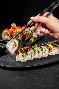 Female hand using traditional Japanese chopsticks to pick up appetizing sushi roll filled with cream cheese, tomato and shrimp topped with wakame seaweed, tobiko and unagi sauce from slate plate