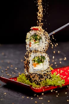 Action shot of toasted sesame seeds being sprinkled on vegetarian sushi roll with cream cheese, carrots, hiyashi wakame, cucumber and chili pepper stacked on red plate against black background