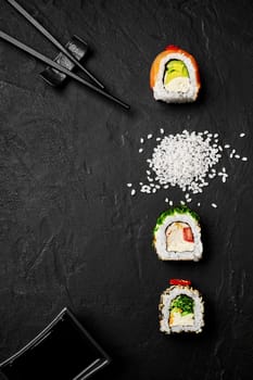 Creative arrangement of three various rolls with salmon, shrimp and vegetables accompanied by chopsticks, soy sauce dish, and scattered sushi rice on textured black slate background. Japanese cuisine