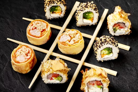 Playful tic-tac-toe arrangement of chopsticks and sushi rolls with various fillings on dark slate background, combining food and fun