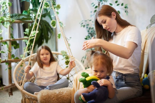 Young loving mom making ponytail to little preschool daughter, adult sister on background in living room. Mother helping child girl with hairstyle