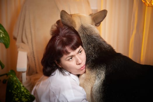 Adult mature woman with big shepherd dog in white shirt. Room with girl and calm cozy evening atmosphere with transparent curtains and soft warm light of lamps. Concept of love for animals, pets