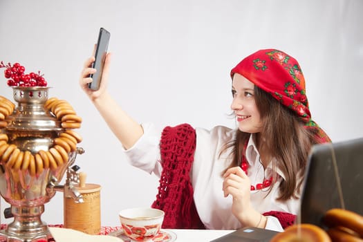 A fashionable modern girl in stylized folk clothes at table with a samovar, bagels and tea takes a selfie on the Orthodox holiday of Maslenitsa and Easter. Funny photo shoot for a young woman