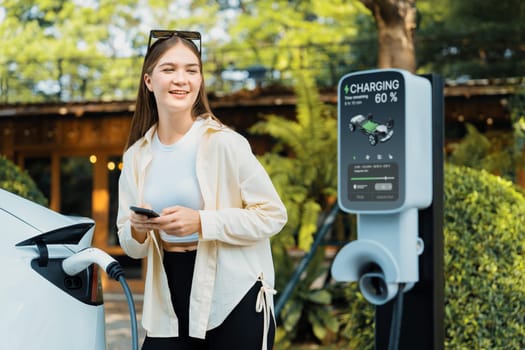 Sustainable urban commute with young woman pay electricity for EV electric car recharging at outdoor cafe in springtime garden, green city sustainability and environmental friendly EV car. Expedient