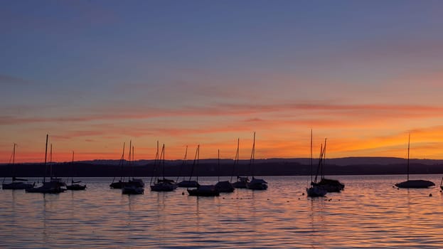 Bodensee Lake Sunrise Panorama. Morning Sunlight Over Tranquil Waters. Witness the mesmerizing dawn over Germany's Bodensee Lake, captured from a boat dock. Embrace the tranquil beauty of the early morning as the sun rises, casting a soft glow on the landscape. The peaceful scene features boats, yachts, and a charming water shack set against a backdrop of a captivating sky. Clouds delicately reflect on the calm water, creating a serene atmosphere. Immerse yourself in the serene beauty of a lakeside sunrise. Explore the harmony of nature, technology, and production as the day unfolds by the lake.