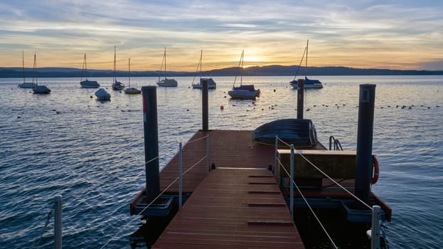 Bodensee Lake Sunrise Panorama. Morning Sunlight Over Tranquil Waters. Witness the mesmerizing dawn over Germany's Bodensee Lake, captured from a boat dock. Embrace the tranquil beauty of the early morning as the sun rises, casting a soft glow on the landscape. The peaceful scene features boats, yachts, and a charming water shack set against a backdrop of a captivating sky. Clouds delicately reflect on the calm water, creating a serene atmosphere. Immerse yourself in the serene beauty of a lakeside sunrise. Explore the harmony of nature, technology, and production as the day unfolds by the lake.