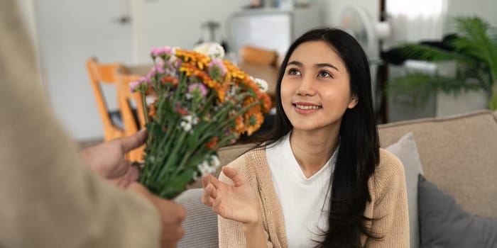 Romantic young asian couple embracing with holding flowers and smiling in living room at home. fall in love. Valentine concept.