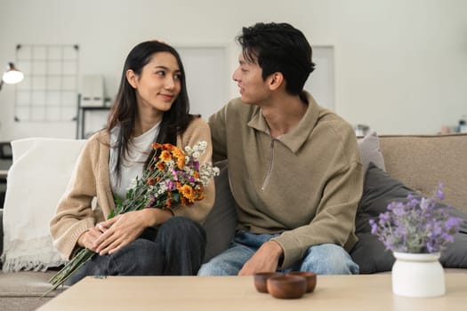 Romantic young asian couple embracing with holding flowers and smiling in living room at home. fall in love. Valentine concept.