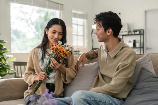 Romantic young asian couple embracing with holding flowers and smiling in living room at home. fall in love. Valentine concept.