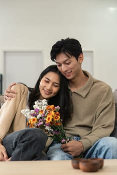 Romantic young asian couple embracing with holding flowers and smiling in living room at home. fall in love. Valentine concept.