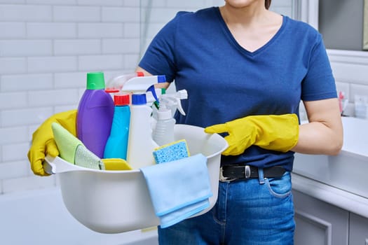 Woman in gloves with basin of detergents in bathroom. Female preparing for routine house cleaning, housecleaning service worker posing at workplace. Housekeeping, housework, cleaning concept