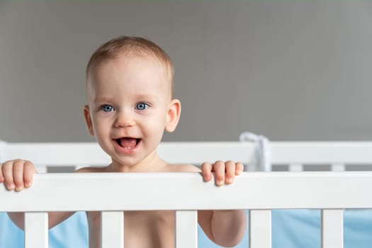 Baby girl with blue eyes standing in crib holding rail and smiling