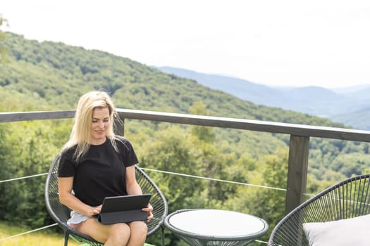 Cheerful girl making video call online on digital tablet. Positive woman talking with friend during video chat on digital tablet. Smiling female hiker sitting on the terrace with mountains on background.