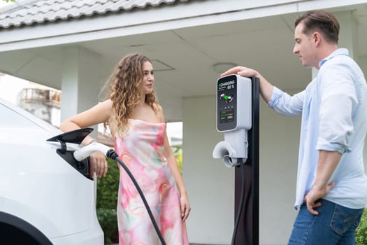 Happy and lovely couple with eco-friendly conscious recharging electric vehicle from EV home charging station. EV car technology utilized for residential home to future sustainability. Synchronos