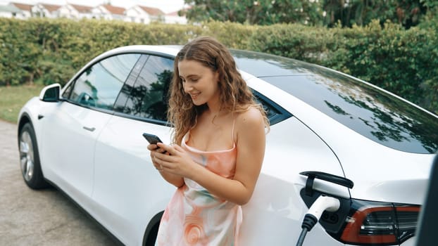 Young woman use smartphone to pay for electricity at public EV car charging station in nature. Modern environmental and sustainable automobile transportation lifestyle with EV vehicle. Synchronos