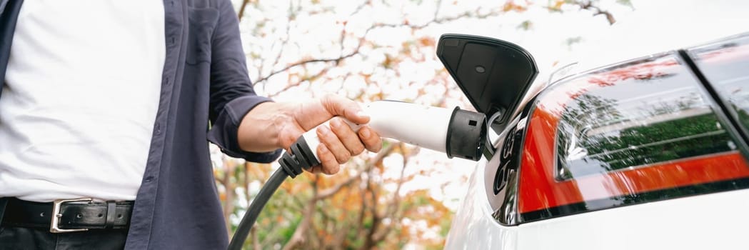 Panorama banner man recharging battery for electric car during autumnal road trip travel EV car in autumnal forest. Eco friendly travel on vacation during autumn with electric vehicle. Exalt