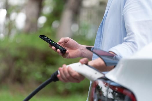 Modern eco-friendly man recharging electric vehicle from EV charging station, using Innovative EV technology utilization for tracking energy usage to optimize battery charging on smartphone.Synchronos