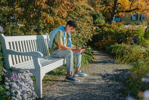 a teenager sits on a bench in the park drinks coffee from a thermo mug and looks into a phone. Portrait of handsome cheerful guy sitting on bench fresh air using device browsing media smm drinking latte urban outside outdoor.