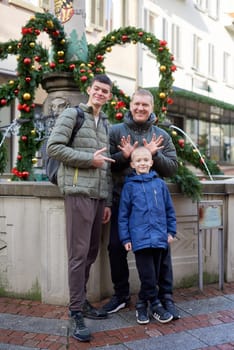 Joyful Family Portrait: Father and Two Sons by Festive Vintage Fountain. Capture the essence of familial happiness with this heartwarming image featuring a handsome father with his two sons standing against the backdrop of a festively decorated vintage fountain. The image beautifully blends the joy of the holiday season with the timeless charm of family togetherness. The father is 44 years old, the elder son is 17, and the younger one is 8, creating a memorable and multigenerational festive scene.