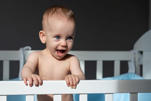 Baby girl with blue eyes standing in crib holding rail and smiling